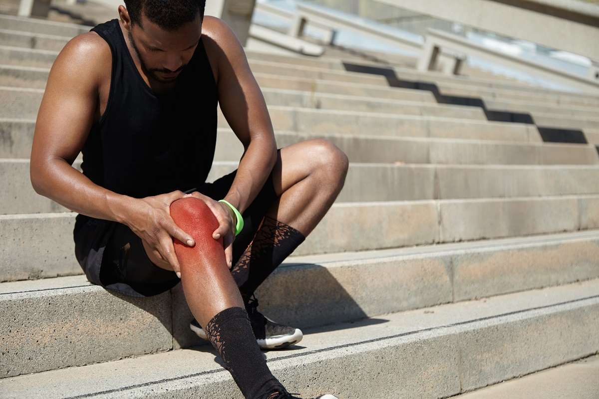 Чернокож мъж в спортен екип, който е седнал на стълби и държи възпаленото си коляно.