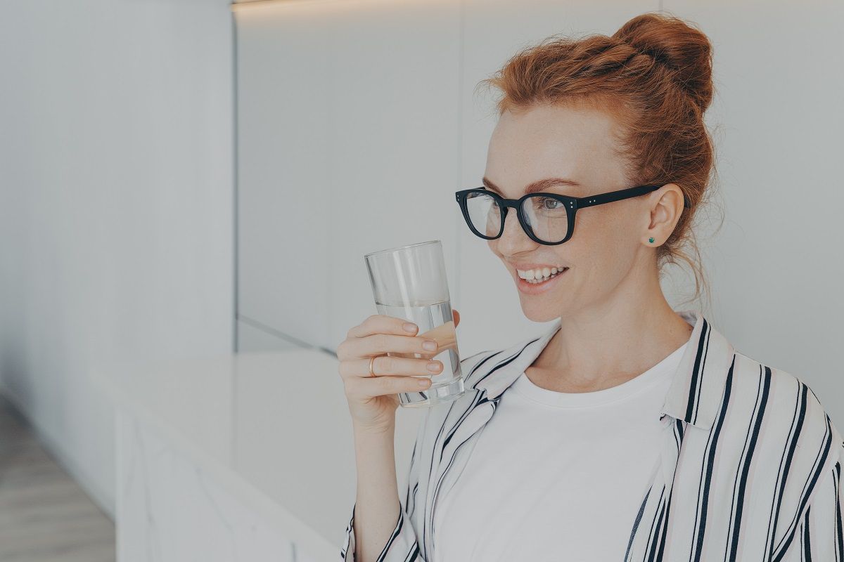 Усмихната жена с червена коса и очила държи чаша с вода